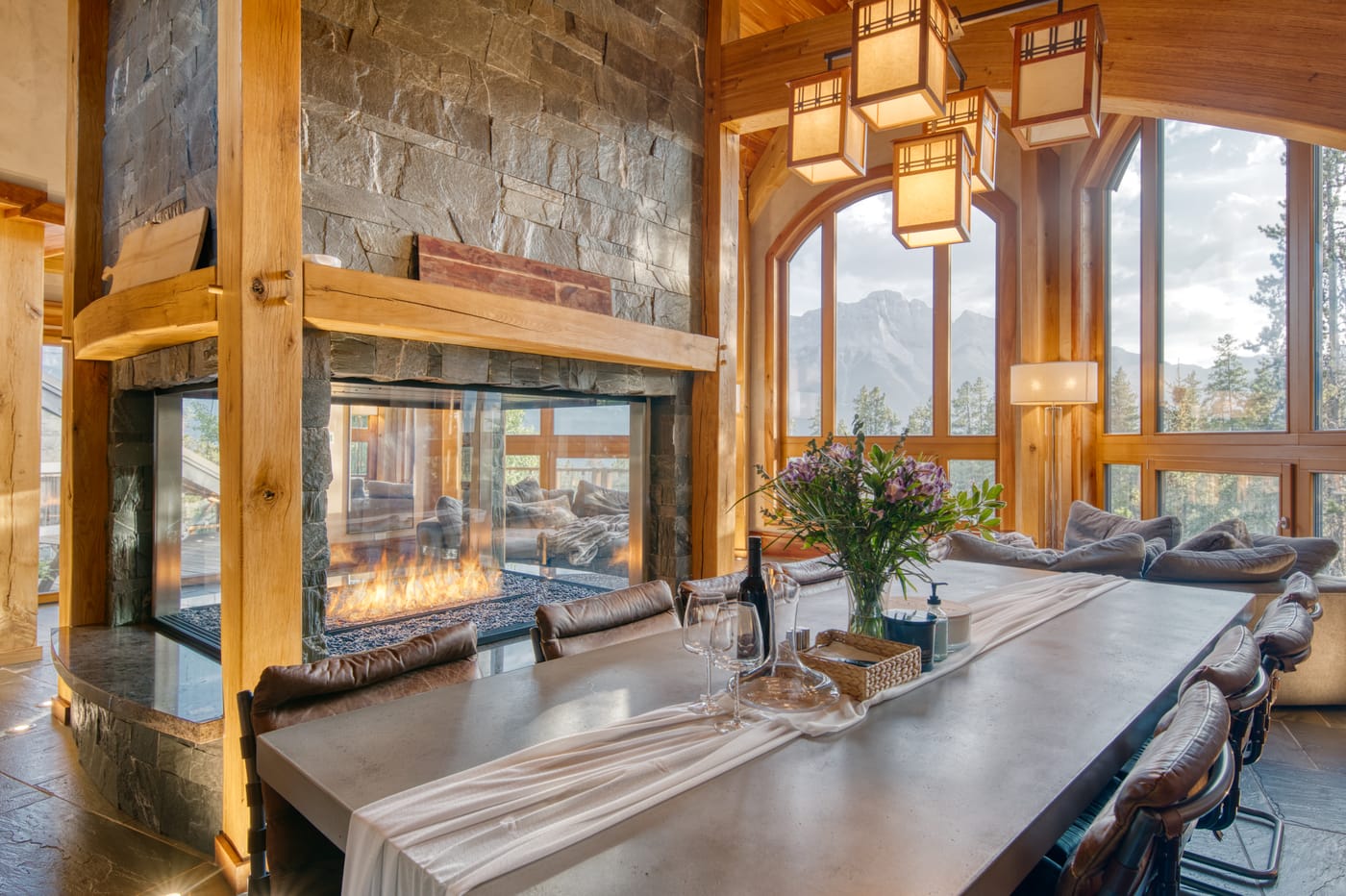 Dining area with a central stone fireplace, timber ceilings, and arched windows onto the mountains